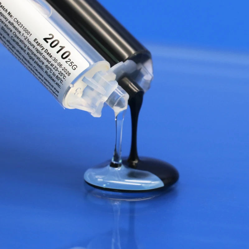 A syringe-style tube of black liquid is tilted over a small clear dish on a bright blue background, with the liquid dripping into the dish. The tube label shows text including &